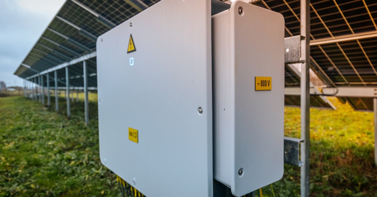 A white solar inverter box mounted under rows of panels in a grassy field with yellow warning labels and cables.
