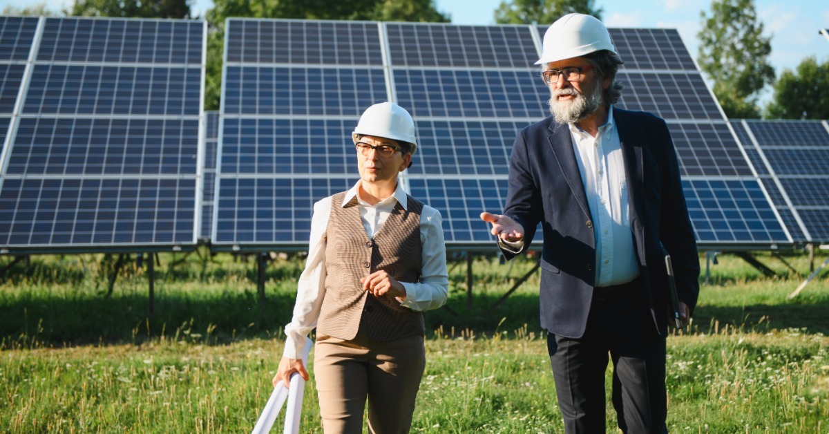 Two people in hard hats walking through grass in front of solar panels, one carrying rolled documents.