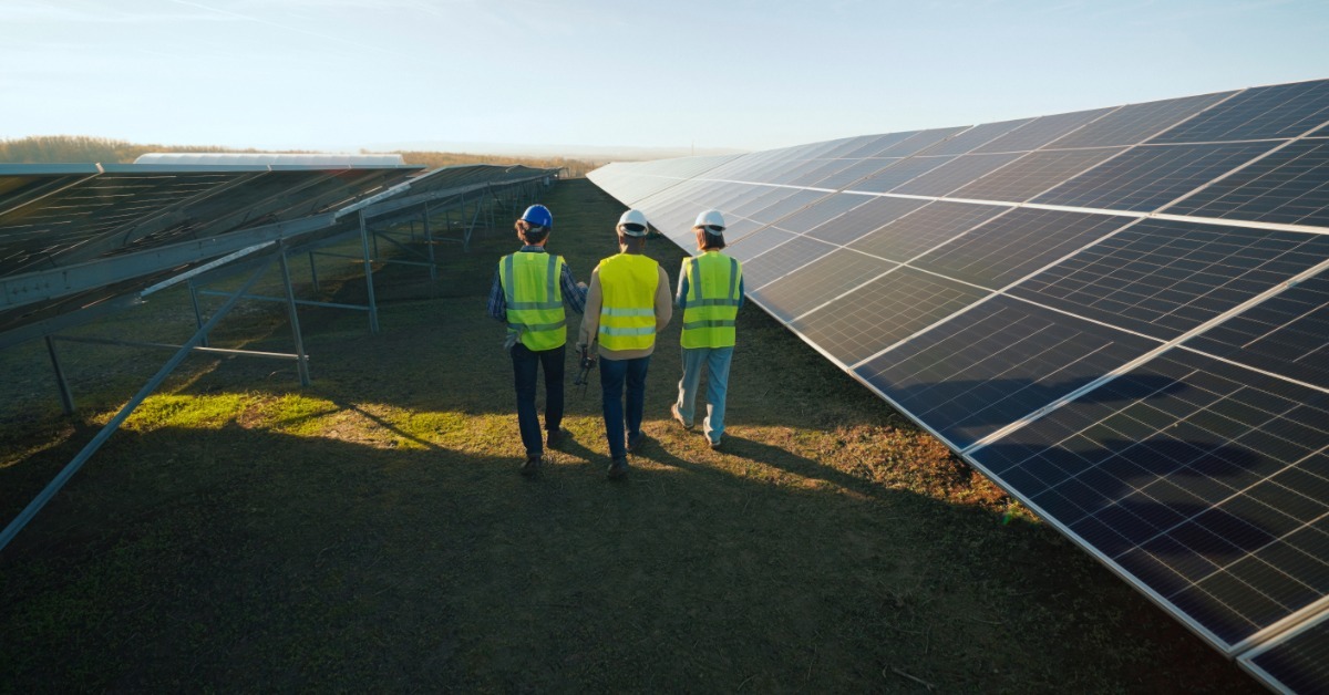 Three people in hard hats and safety vests walking between rows of solar panels under a bright blue sky.