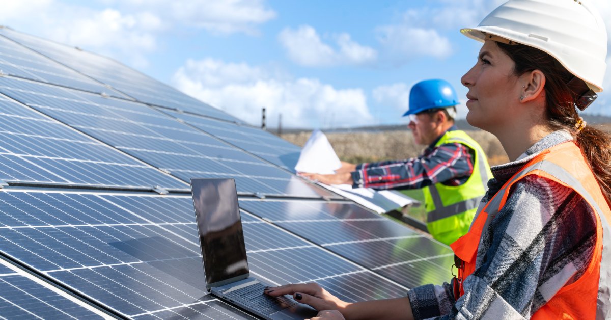 Two engineers in hard hats working beside solar panels, with one using a laptop and another holding papers.