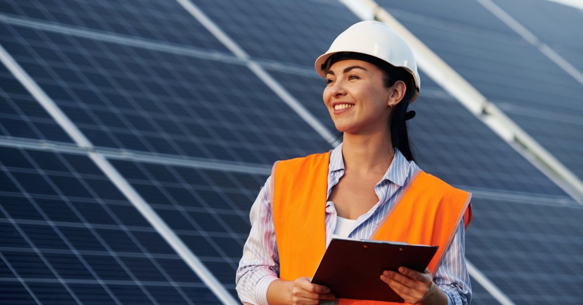 A person wearing a white hard hat and orange safety vest holding a clipboard beside rows of solar panels.