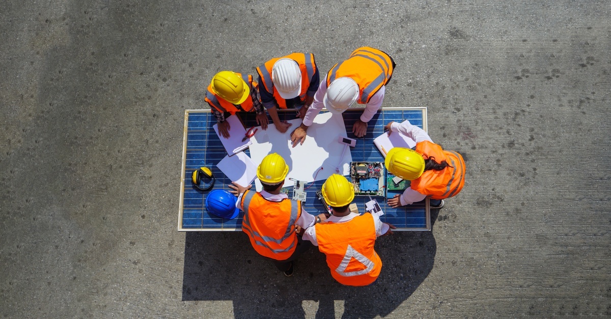 A group of people wearing safety gear huddled around a solar panel with several documents scattered across its surface.