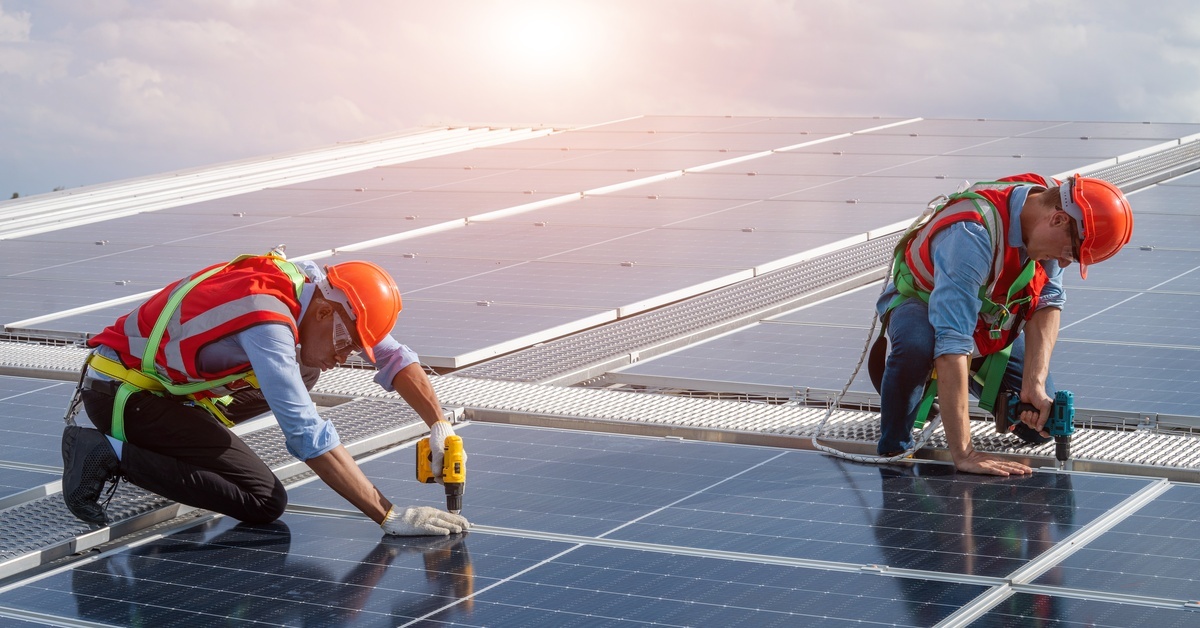  Two workers are installing a solar panel into the frame. They are both wearing safety gear and kneeling on the panel.