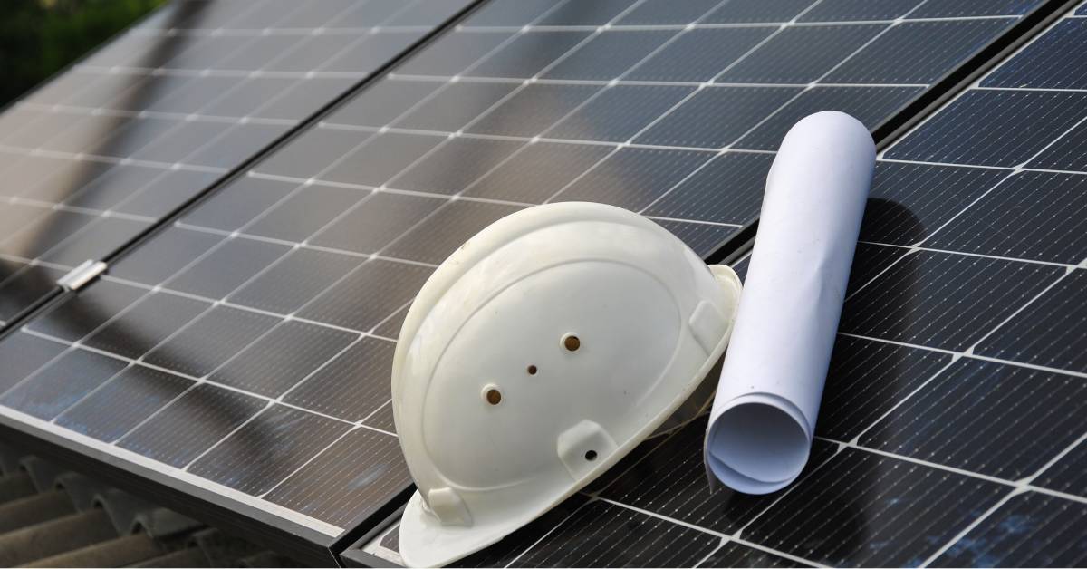 A worker's helmet and a blueprint sit at the edge of a solar panel grid. The blueprint is rolled into a paper roll.