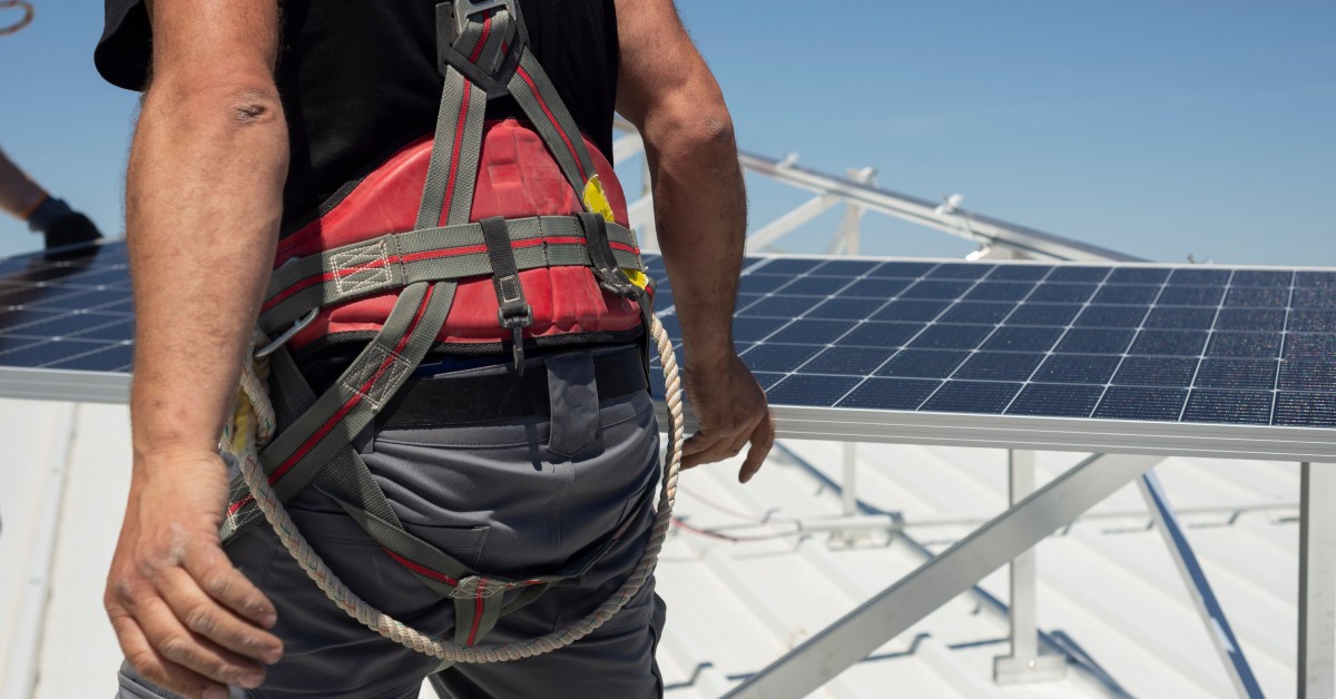 A technician on a roof with solar panels installed. He is wearing a harness while he is walking on the roof.