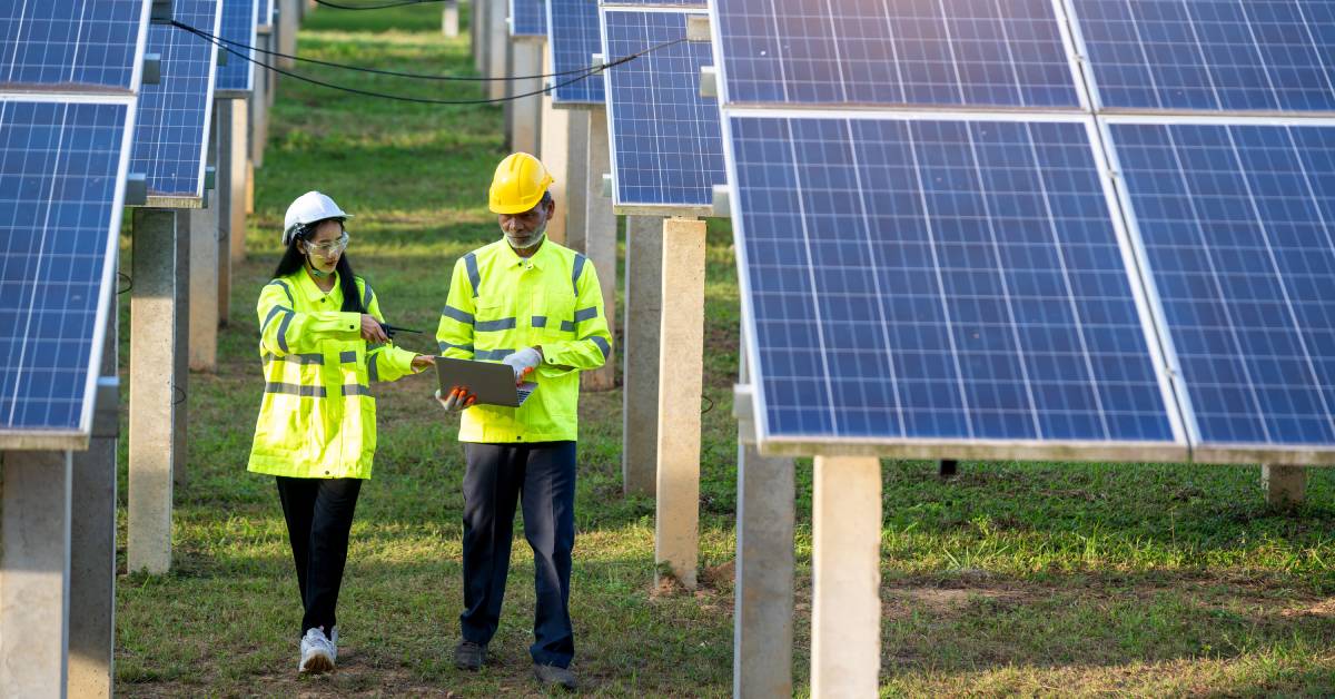 Two maintenance workers in reflective vests and hard hats walking down the aisle of a solar farm. One is using a laptop.