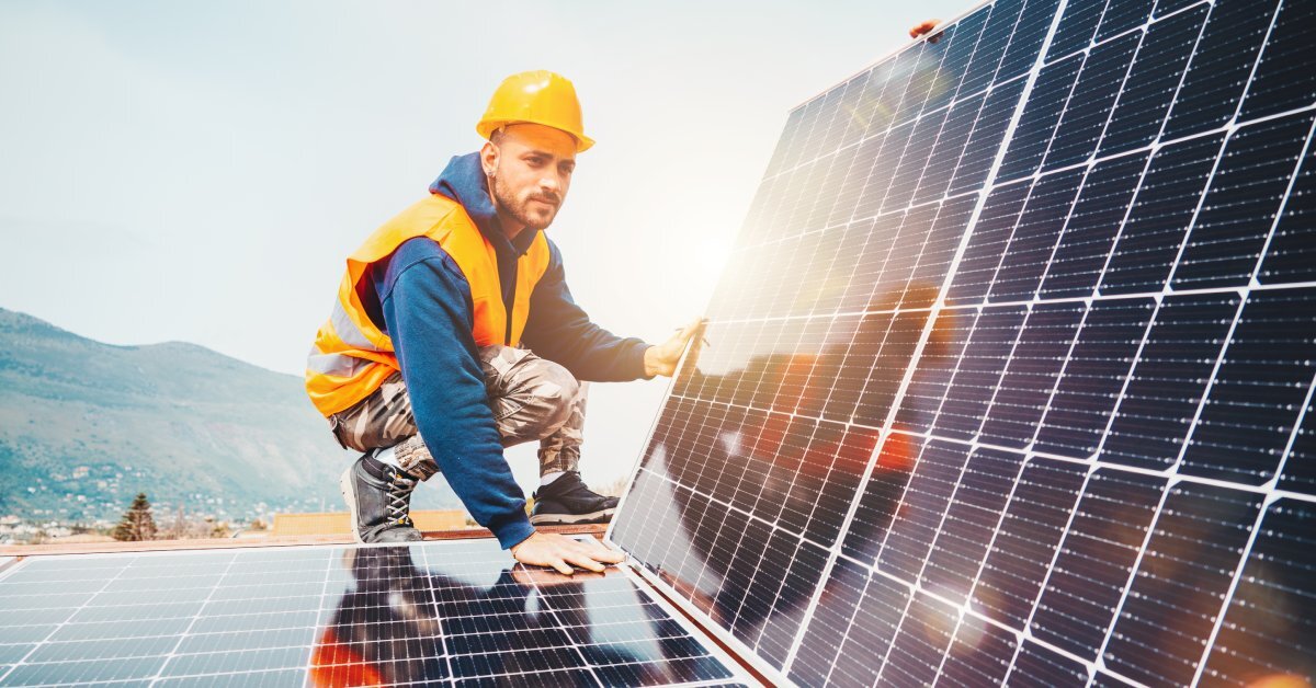 A solar engineer wearing an orange hard hat. He is holding a solar panel at an angle with the ground as leverage.