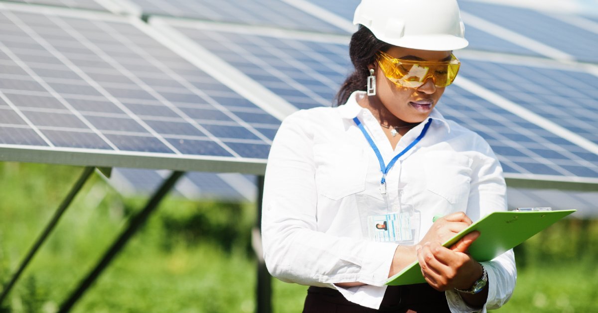 A woman wearing a white hard hat and holding a green clipboard. She is standing in front of a solar grid.