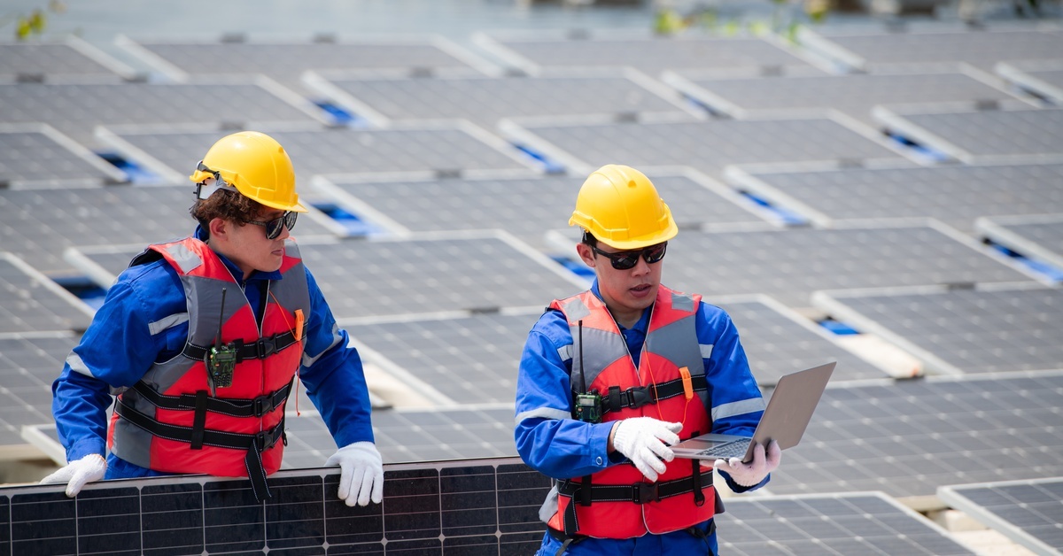 Two solar grid worker shown among solar panels. One holds a laptop and the other looks at the screen while holding a solar panel.