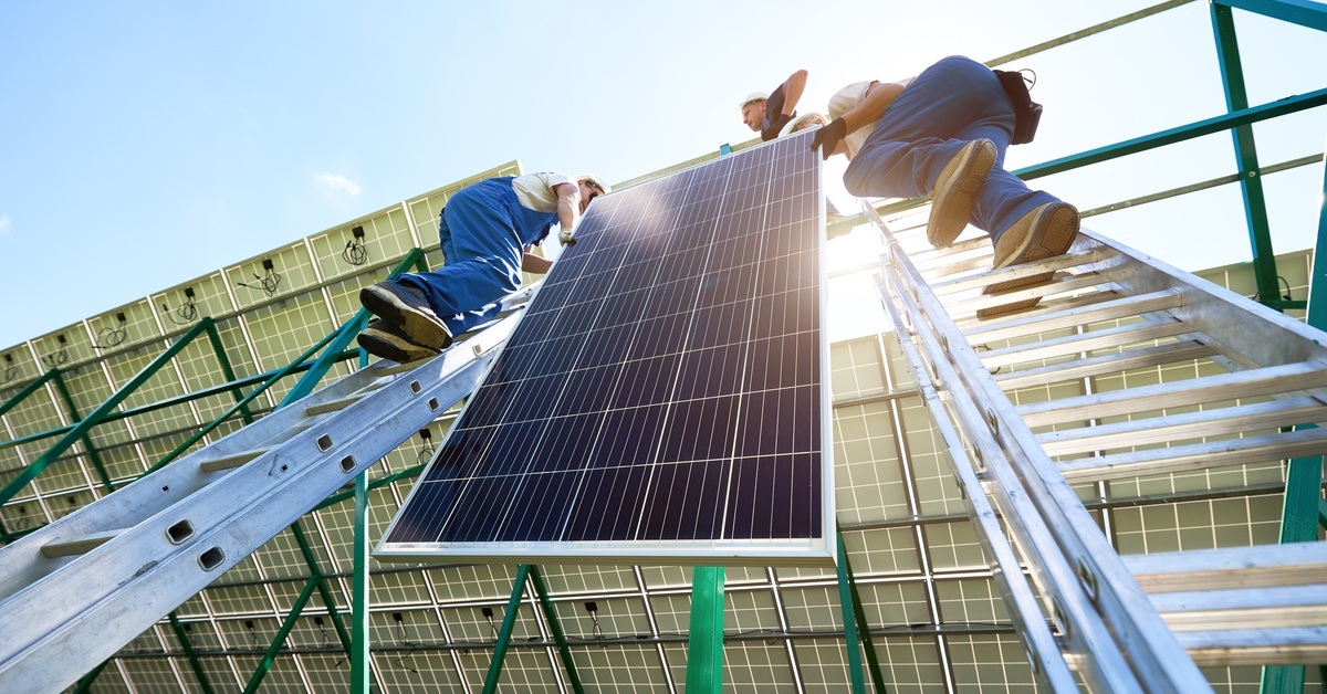 Two people standing at the top of an industrial ladder propped against a structure. They are holding a solar panel.