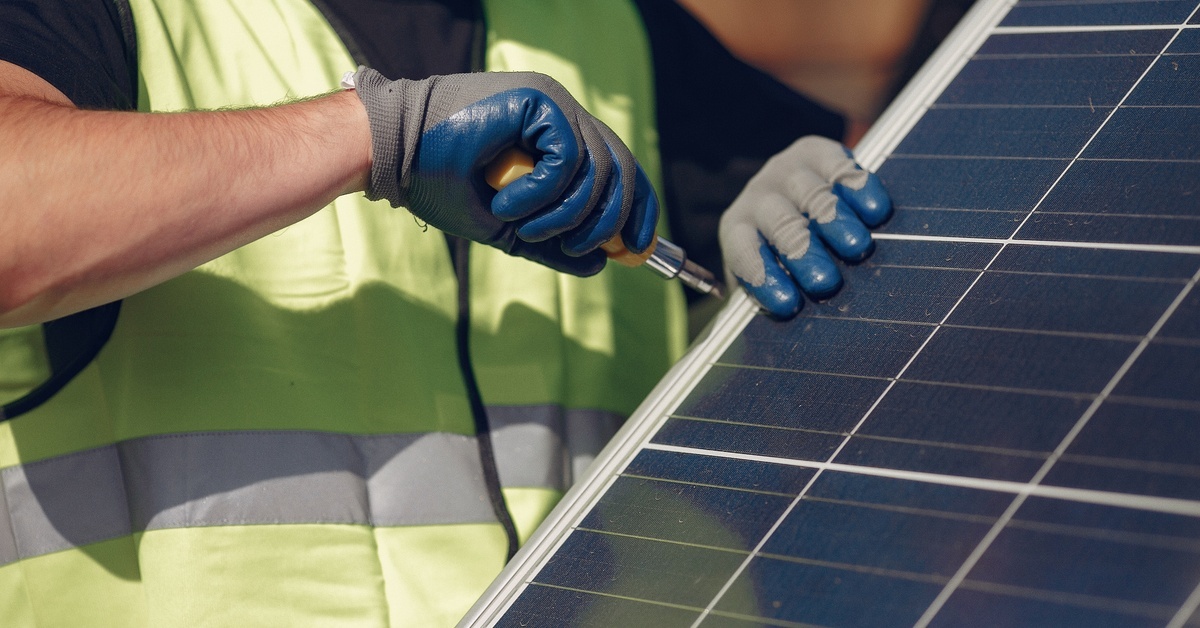 A person using a screwdriver at the edge of a solar panel. They are wearing protective gloves and a reflective vest.