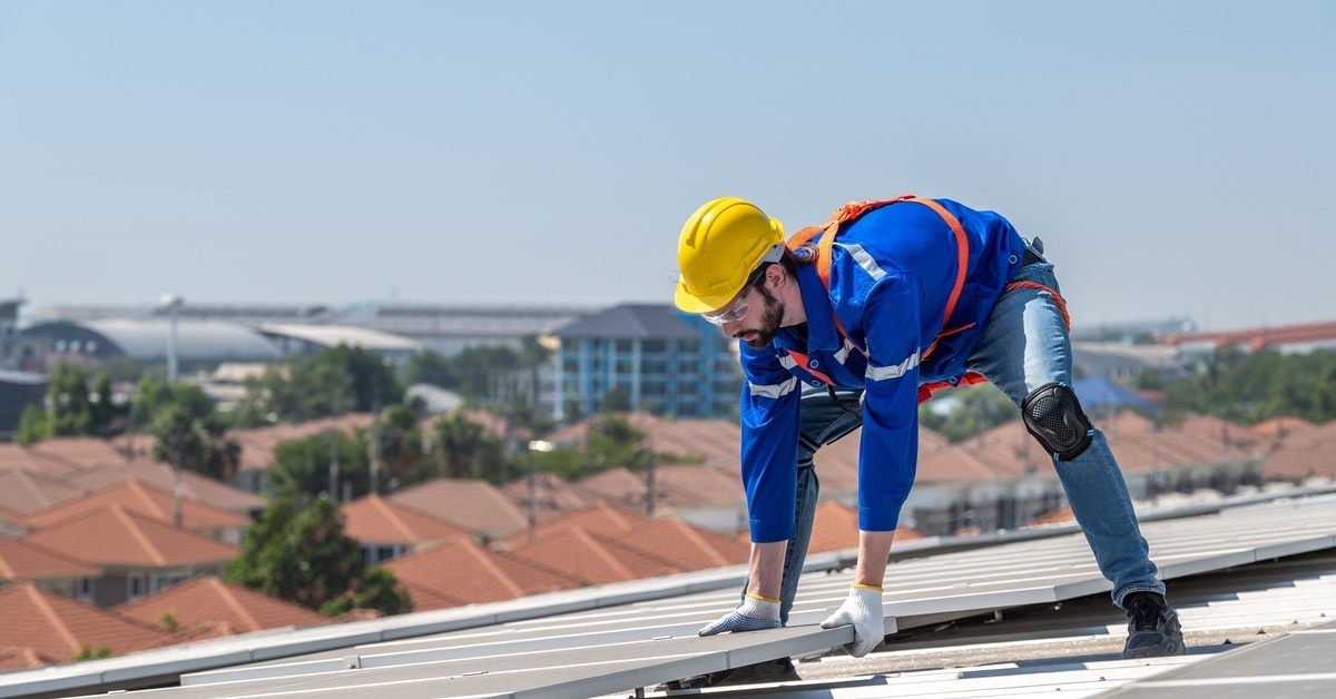 A solar engineer placing a solar panel down on a roof. He is wearing a blue reflective jacket and a yellow hard hat.