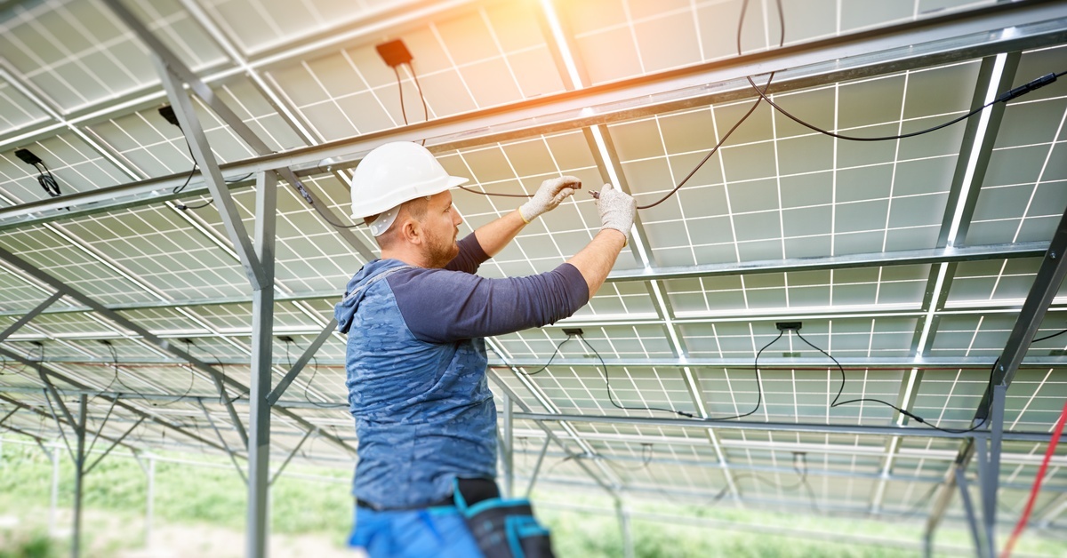 A solar electrician connects two cables together underneath a solar panel grid while wearing gloves and a hard hat.