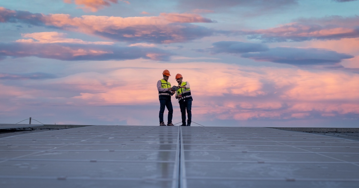 Two men standing in front of a solar panel grid wearing blue hard hats. One of them is showing the other his phone.
