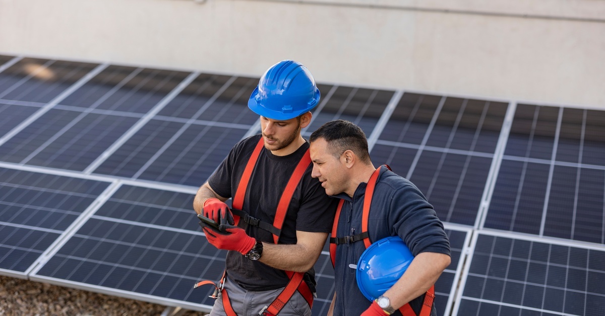 Two men standing in front of a solar panel grid wearing blue hard hats. One of them is showing the other his phone.