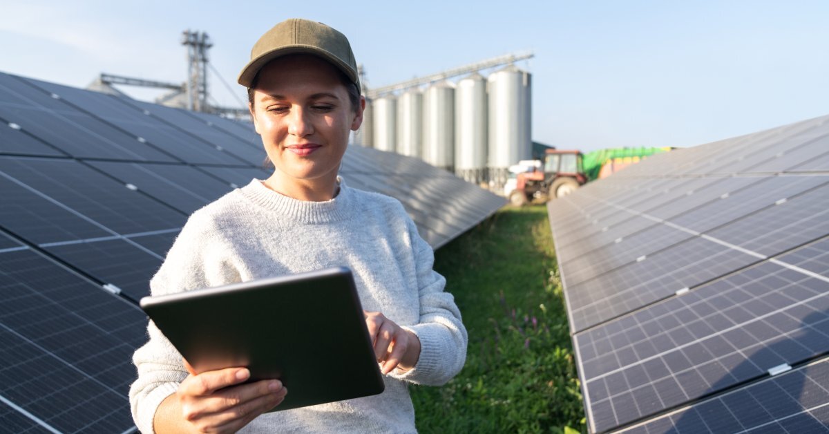 A female farmer walking between large solar panels while looking at an iPad. There are farm silos behind the panels.
