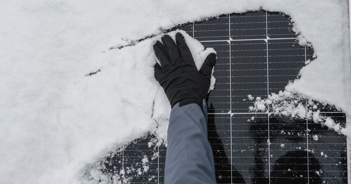 A close-up of a person wearing a long-sleeved jacket and black gloves wiping a pile of snow off a solar panel.