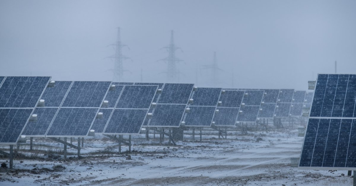 Solar panels with snow falling around them and sticking to the ground. Power lines are visible in the distance.