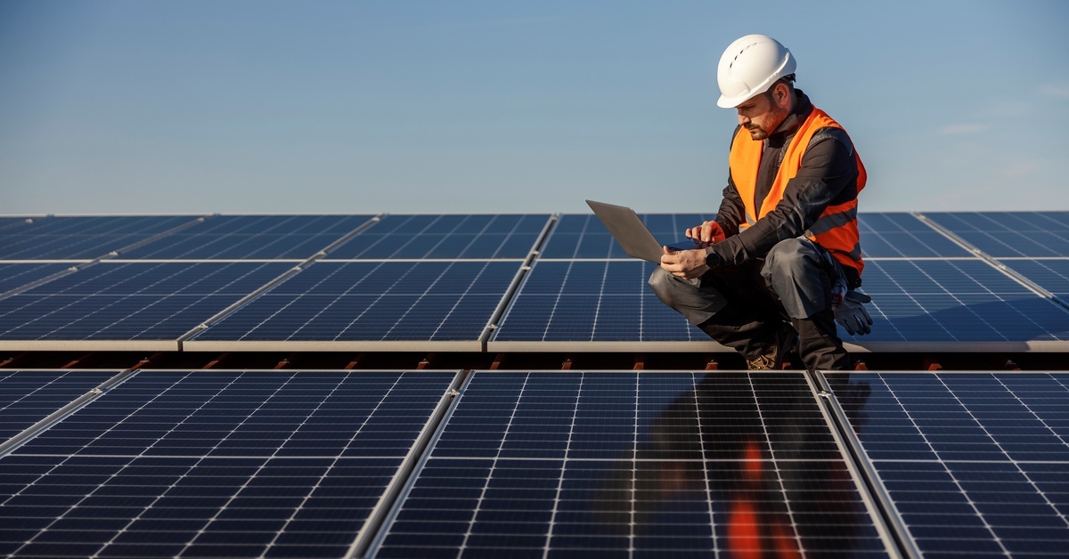 A worker holding a laptop looking at the device while standing in the middle of a large solar panel.