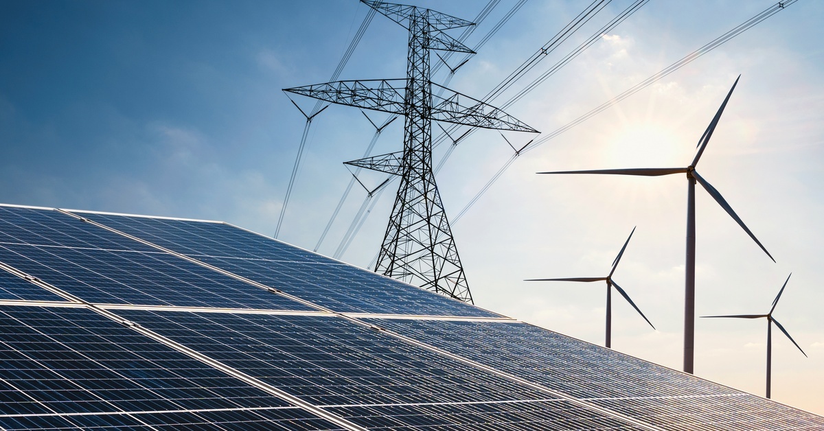 A solar panel, with an electrical tower and windmills in the distance behind it, a blue sky behind the power sources.
