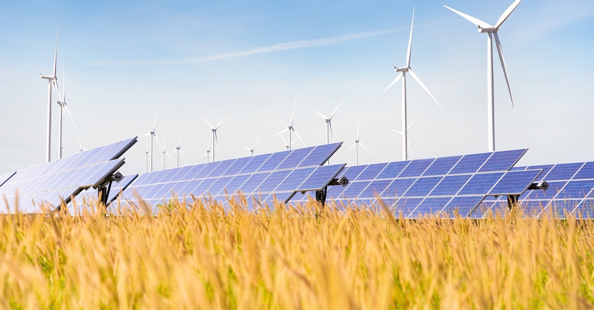 In a field of grass, solar panels are on the ground, and windmills are high above the field to generate energy.