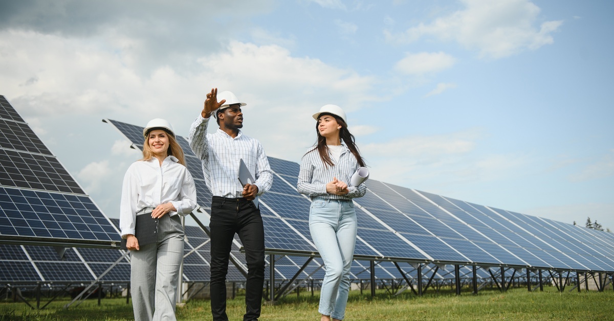 A man walking with two women through a field of solar panels as they discuss something. They are all wearing hard hats.