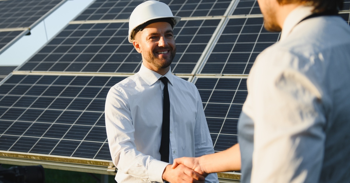 Two men in business attire shake hands while standing in front of large solar panels. One man is wearing a hard hat.
