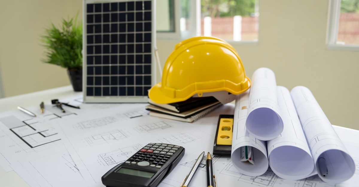 A table filled with supplies for a solar energy project. A hard hat, blueprints, and a calculator are scattered on the table.