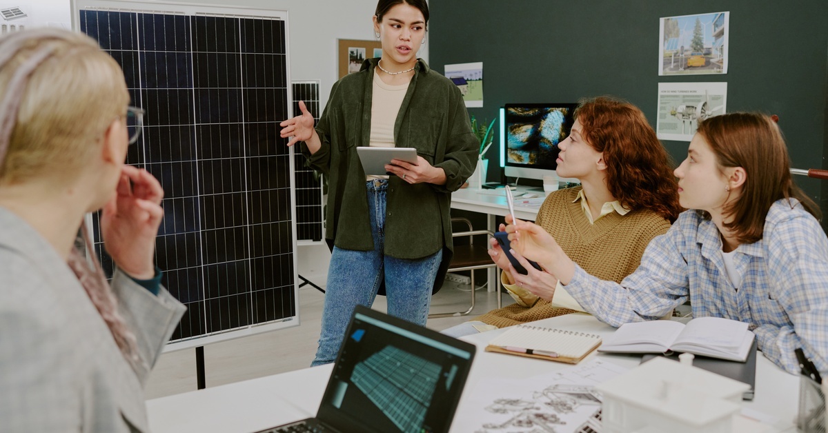 A woman standing in front of a table of people giving a presentation. She is presenting a renewable energy proposal.