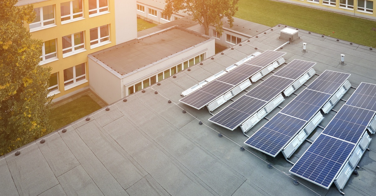 The roof of a school with multiple rows of solar panels placed on it. There are other buildings around the school.