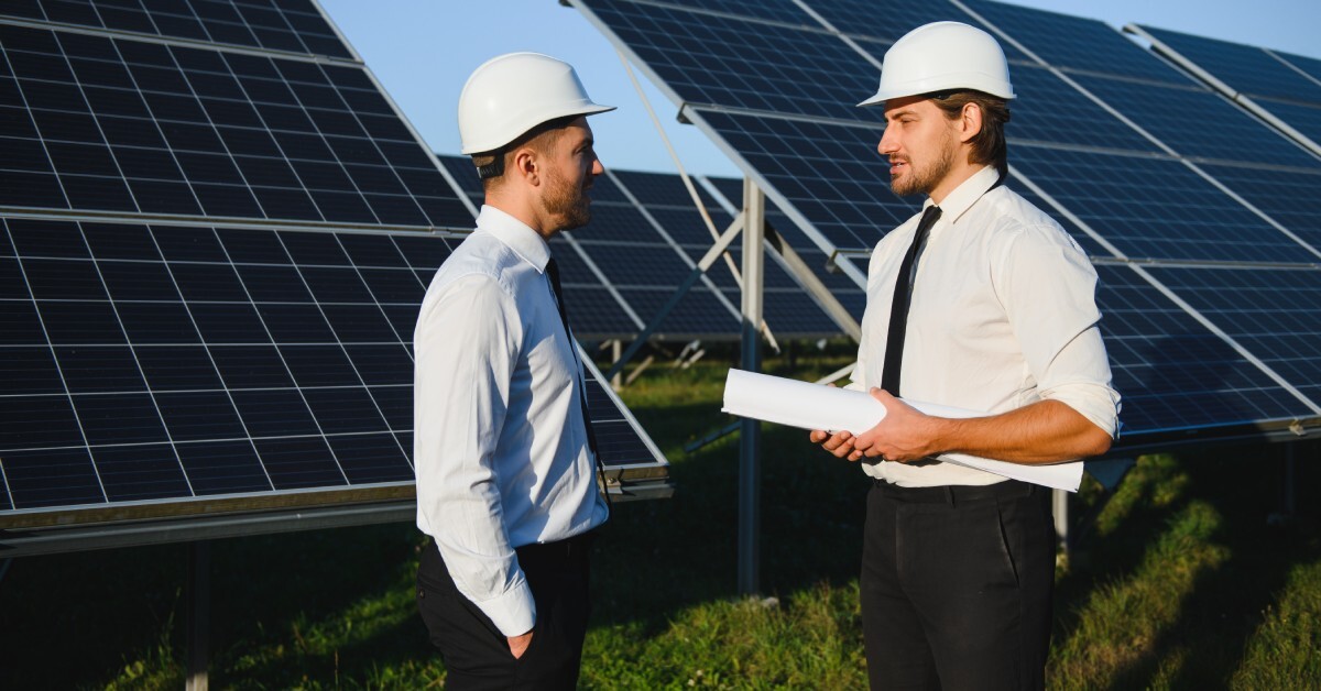 Two businessmen in work attire speaking in front of large solar panels. Both men have safety helmets on.