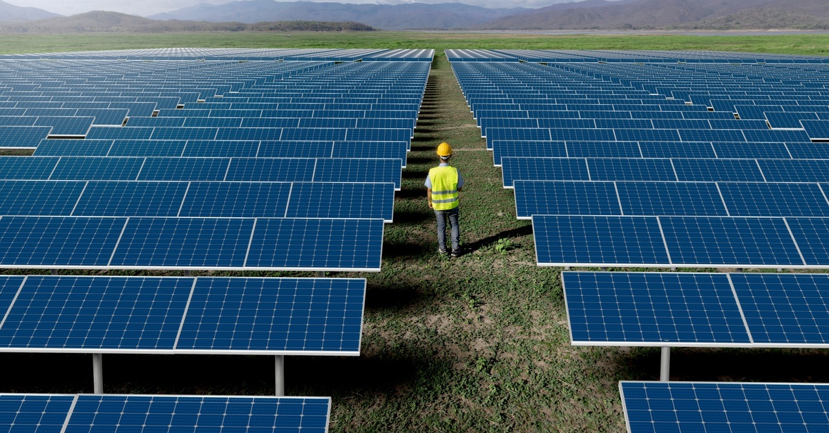 8 Tips for Solar Panel Maintenance and Cleaning An engineer wearing a hard hat and high-visibility vest stands in the middle of a row of solar panels on a solar farm.