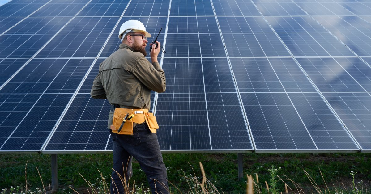 An engineer wearing a protective helmet standing in front of a solar panel. He is talking on the radio.