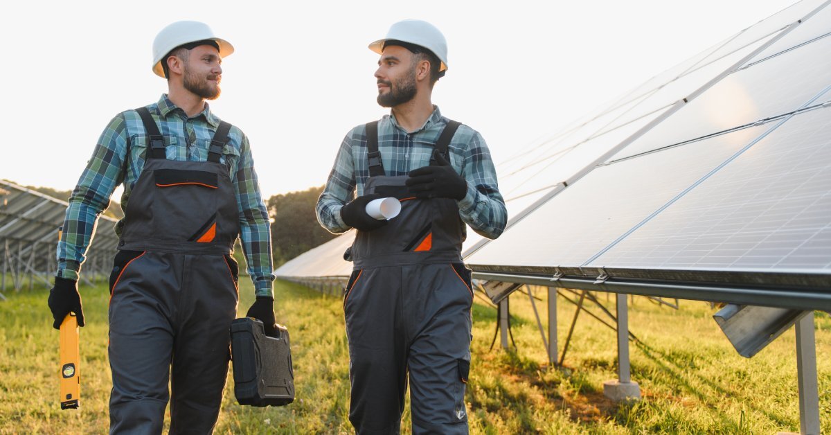 Two technicians walking on a solar farm discussing something. They both have uniforms and hard hats on to stay safe.