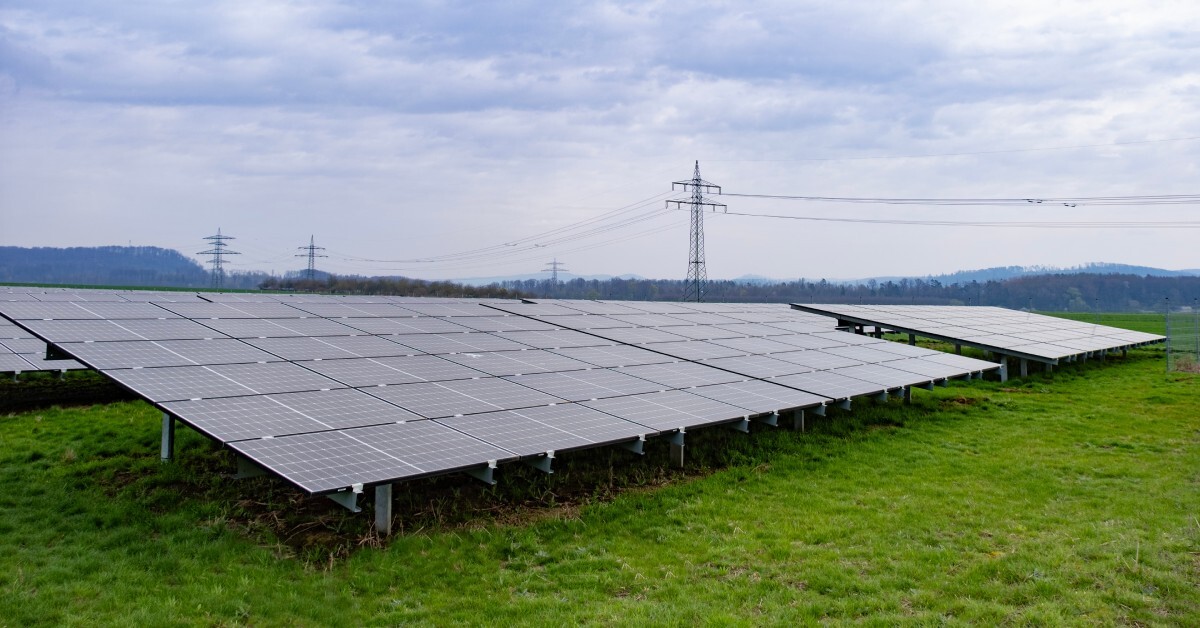 A green field contains rows of solar panels. Large utility poles stand tall in the background against a cloudy sky.