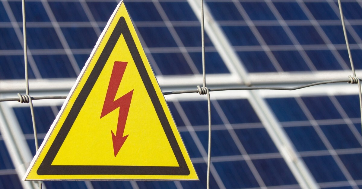 A triangle warning sign indicates there is electrical power behind a fence with multiple solar panels.