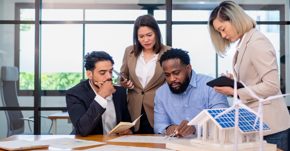 A group of people talk together while looking at research. Models of renewable energy methods sit on a table near them.