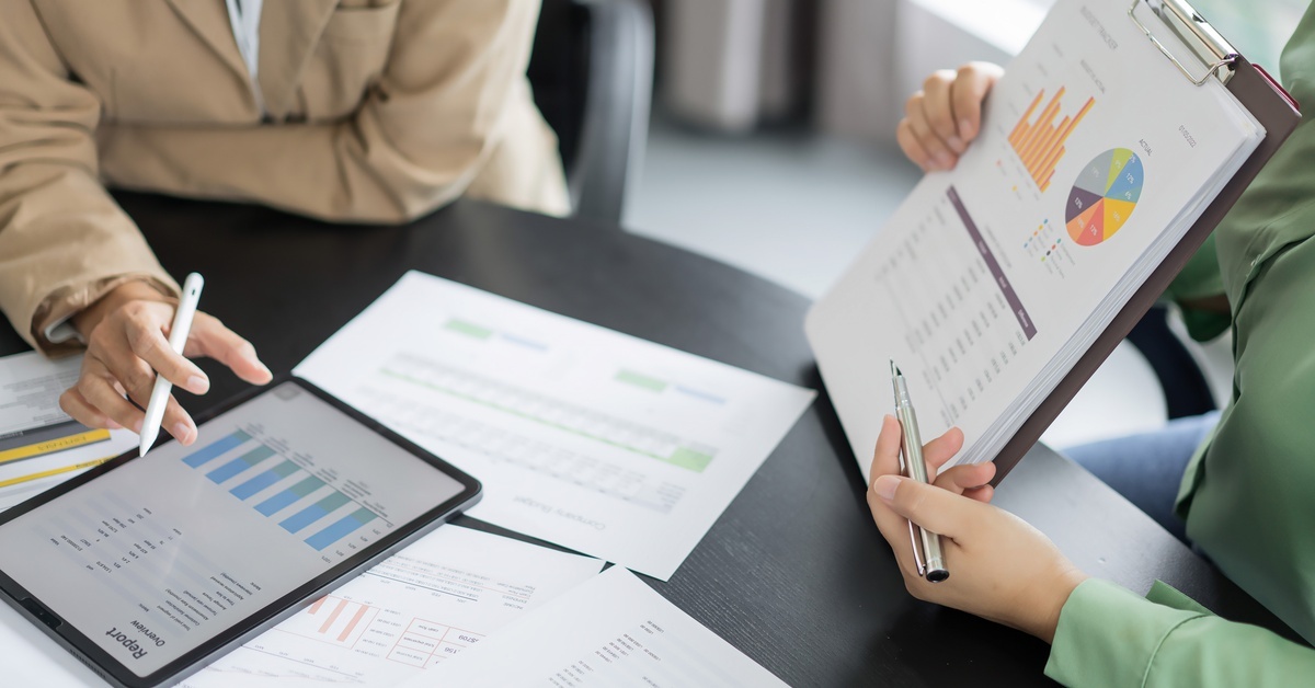 Two people analyze research data on renewable energy. One of them holds paper records, while the other looks at a tablet.