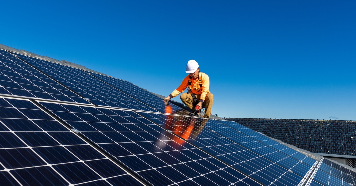A solar panel technician wearing standard safety gear stands next to rows of panels, drilling a new one in.
