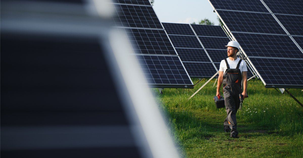 A solar farm worker walking through a field of large solar panels. There is tall grass under the panels.