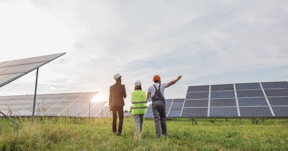 The backs of inspectors and workers as they walk through a field of solar panels. One of them is pointing at something.