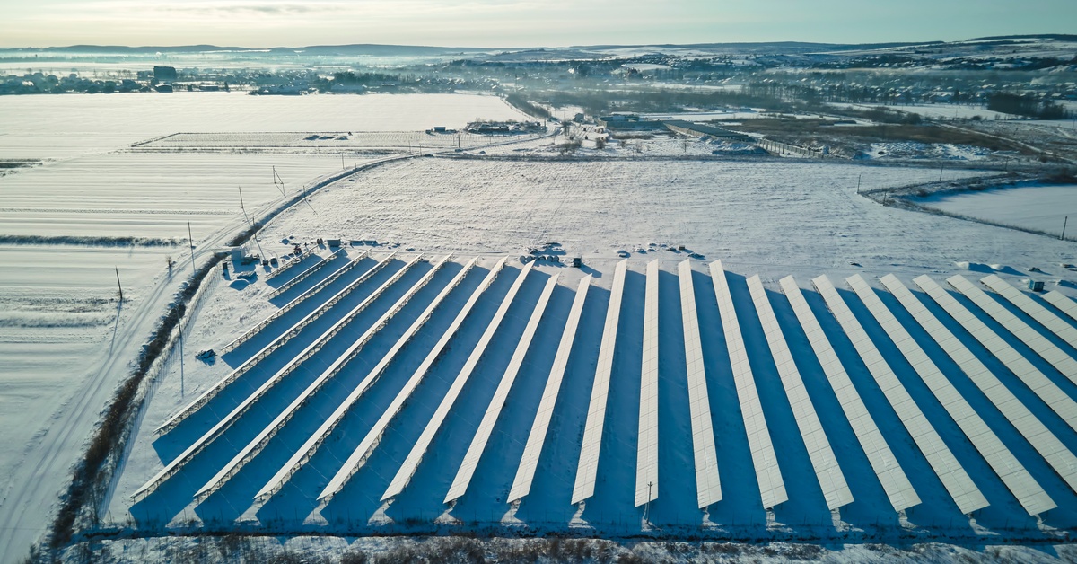 A field partially filled with solar panels covered in a blanket of snow. The panels are fully obscured due to the snow.