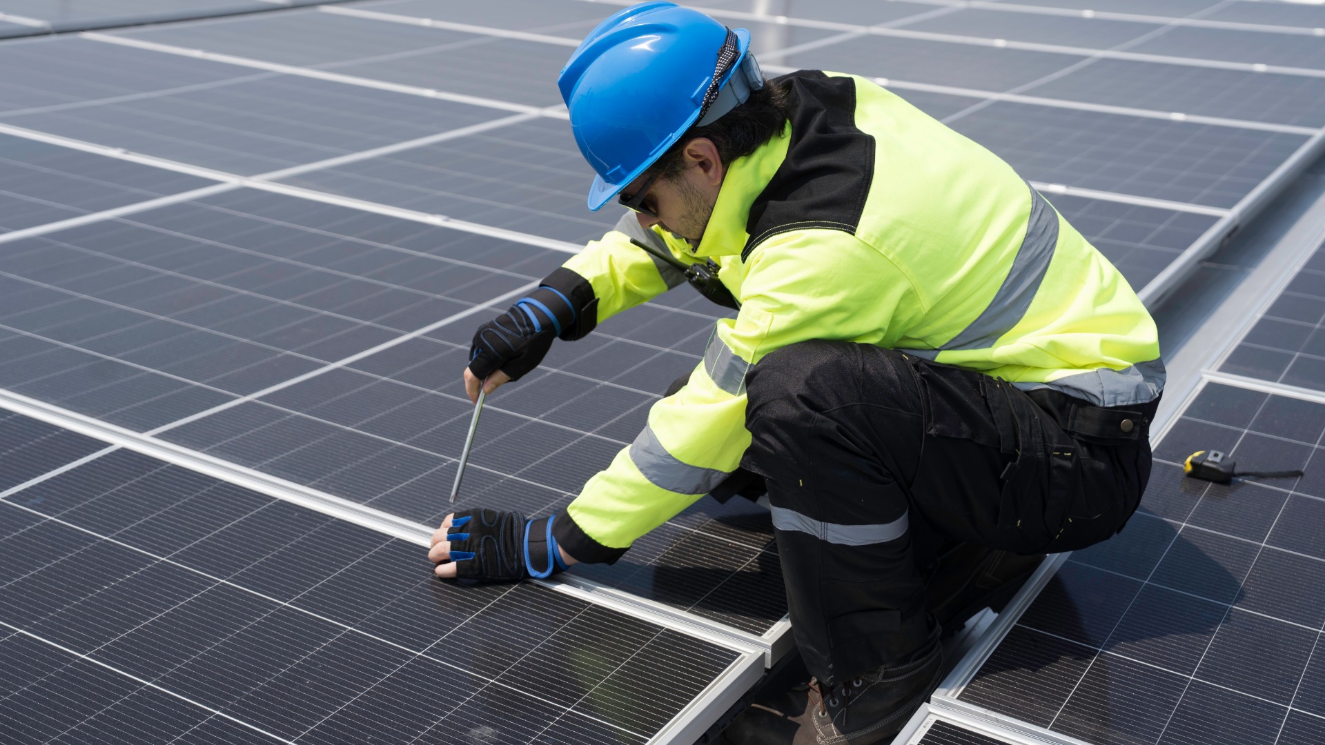An engineer in safety gear is repairing a solar panel. He is crouched next to a row of panels with a screwdriver.