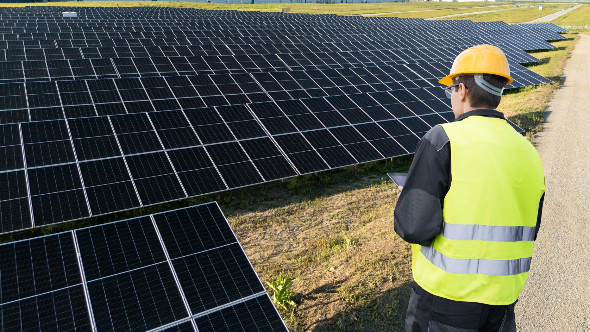 On a sunny day, an engineer with a tablet is looking out at a field of solar panels. He is also wearing a hard hat.