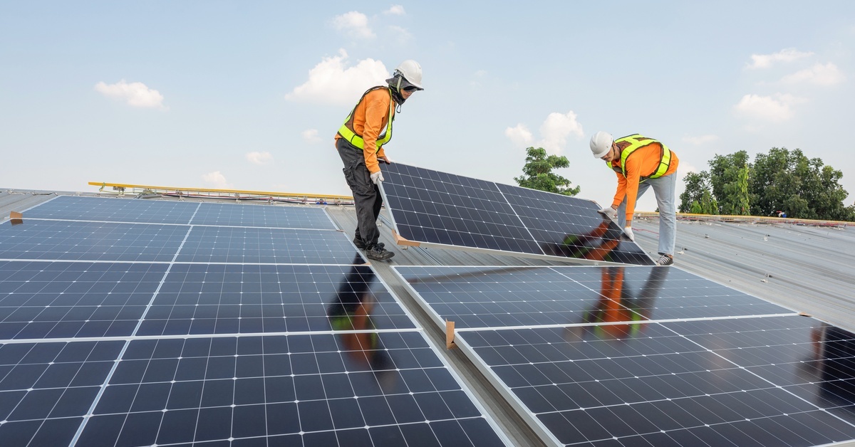 Two technicians installing solar panels on a rooftop. They are both wearing safety gear and hard hats.