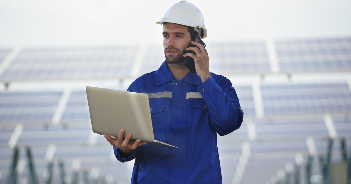 An engineer working on a solar farm with tall panels behind him. He is on the phone and holding a laptop.