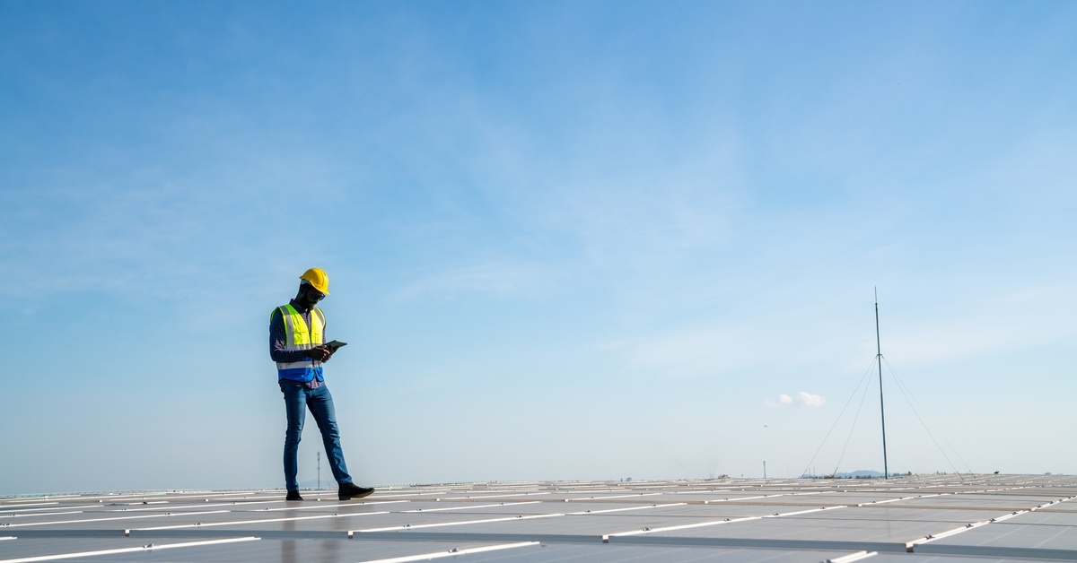 An engineer walking across a large installation of solar panels with a tablet in his hands. The sky above is clear.