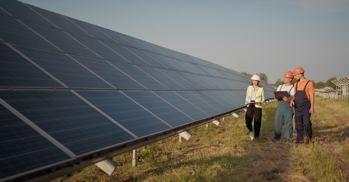 A group of industrial engineers walking next to solar panels and examining them. One of the engineers has a laptop.