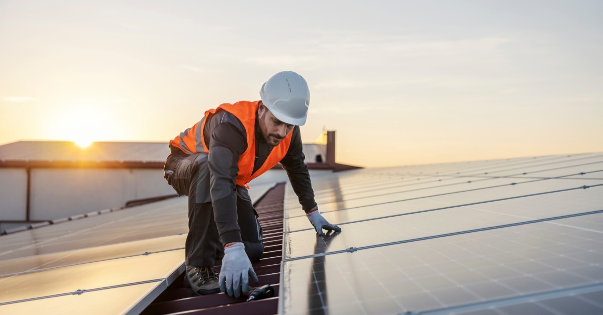 A worker in safety gear with his hands down on a solar panel, working to fix it. He is crouched on a roof.