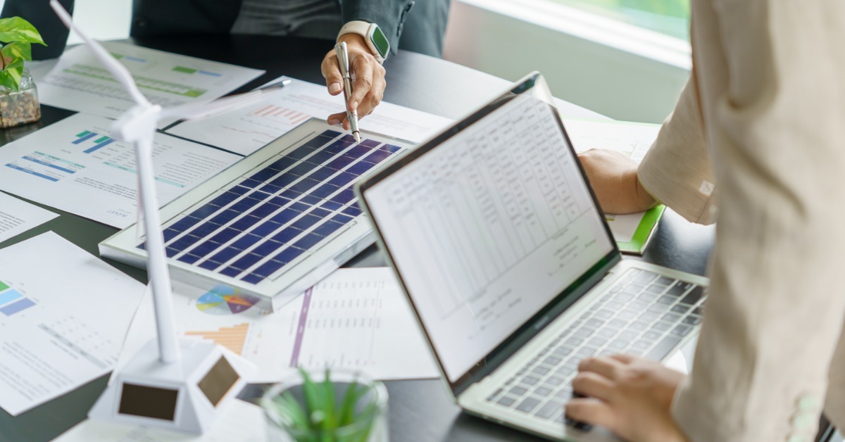 Business people standing over a desk with renewable energy dioramas in front of them. One of them is on a laptop.